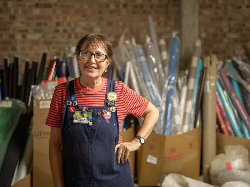 A Scrapstore volunteer in badge-covered dungarees, standing among rolls of paper and card
